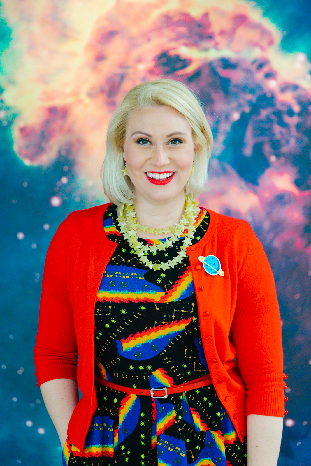 Event Planner Holly Gray wearing bright red cardigan and planet pin stands in front of SKETCH Foundation Air and Space signage featuring image of space