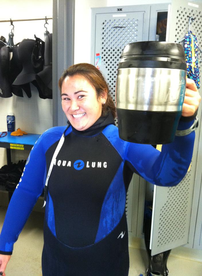 Wearing a wetsuit with her hair pulled back as she stands in the California Science Center's dive locker, Erin Shusterman holds her coffee mug in a gesture that seems to say "cheers" 