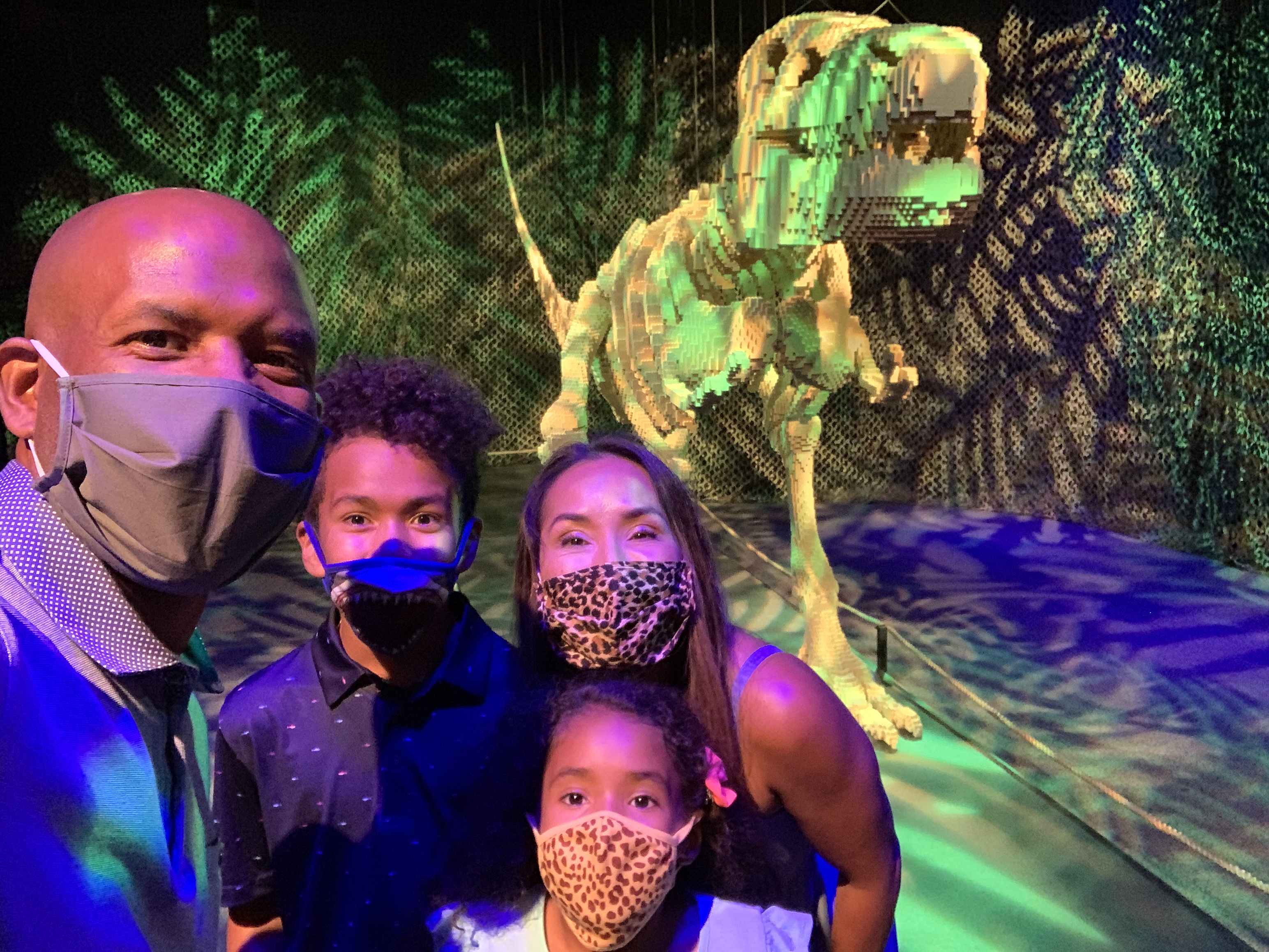 Diverse family with face masks taking a selfie in front of T-Rex sculpture in The Art of the Brick exhibition