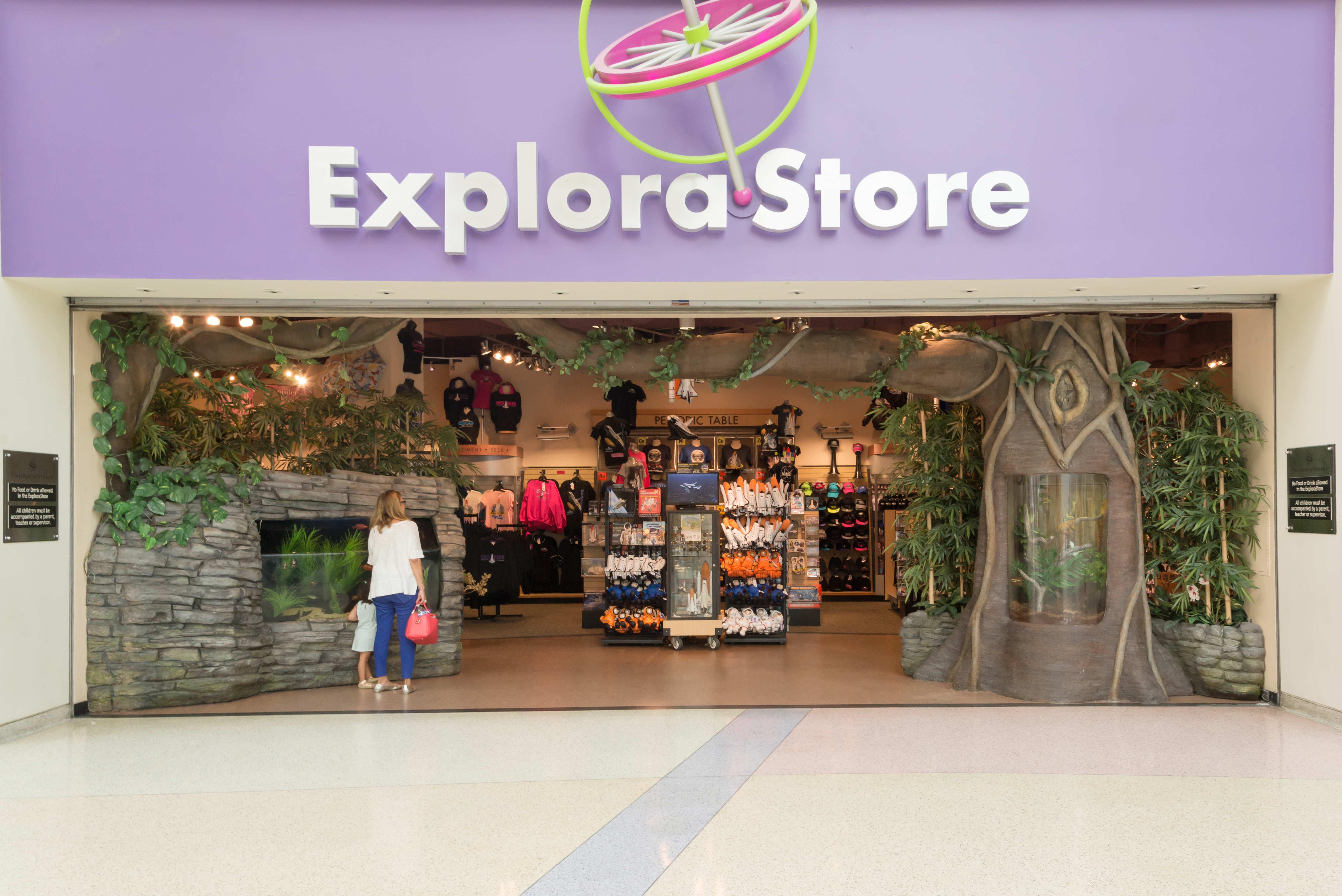 Woman and child stand outside ExploraStore entrance. Upper entry signage reads "Explorastore" and features a large decorative gyroscope. Racks of store merchandise can be seen inside.