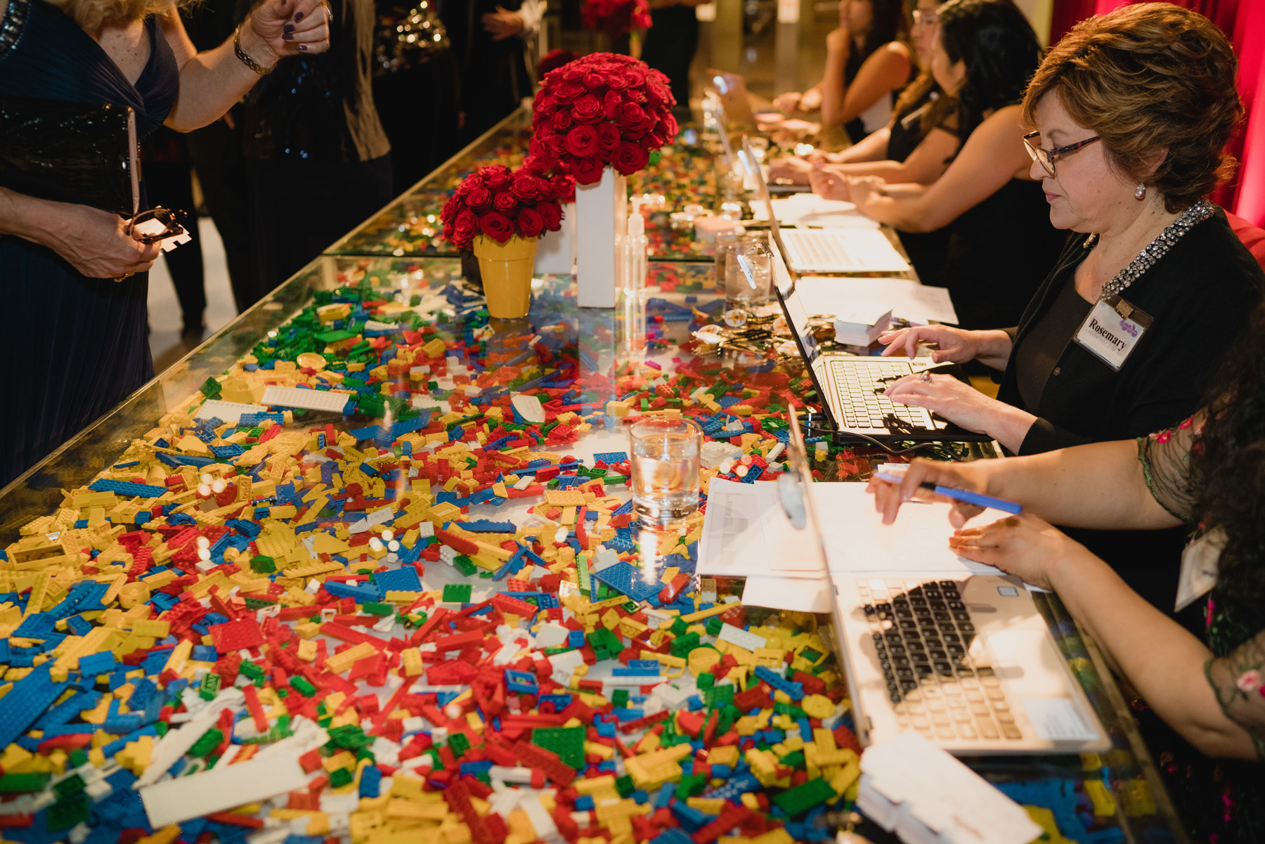 Discovery Ball registration staff working on laptops to check-in guests while sitting at long clear, acrylic table filled with hundreds of LEGO bricks in standard LEGO colors.
