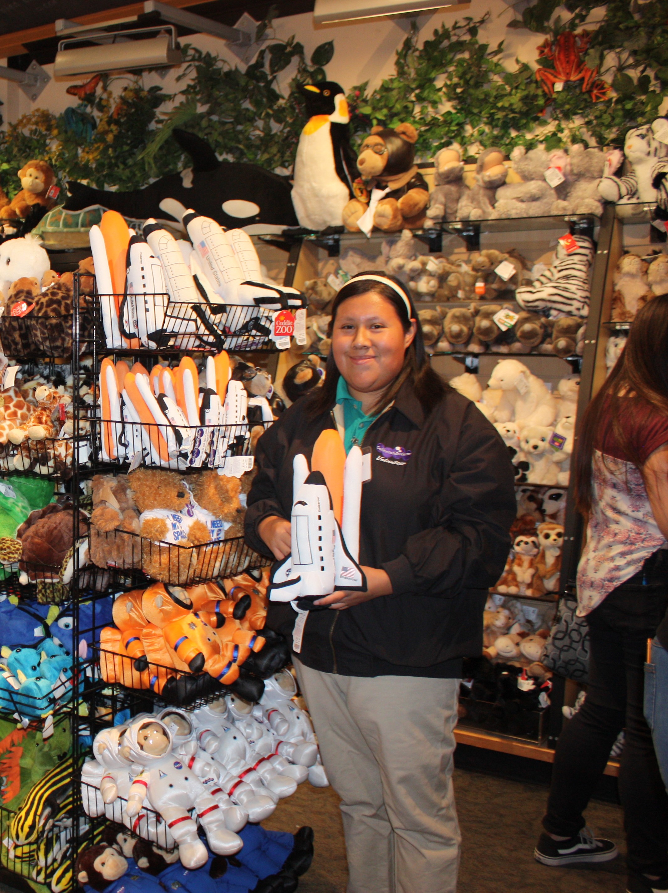 ExploraStore Volunteer holds plush Endeavour Toy next to retail rack of space themed plush toys inside the store.