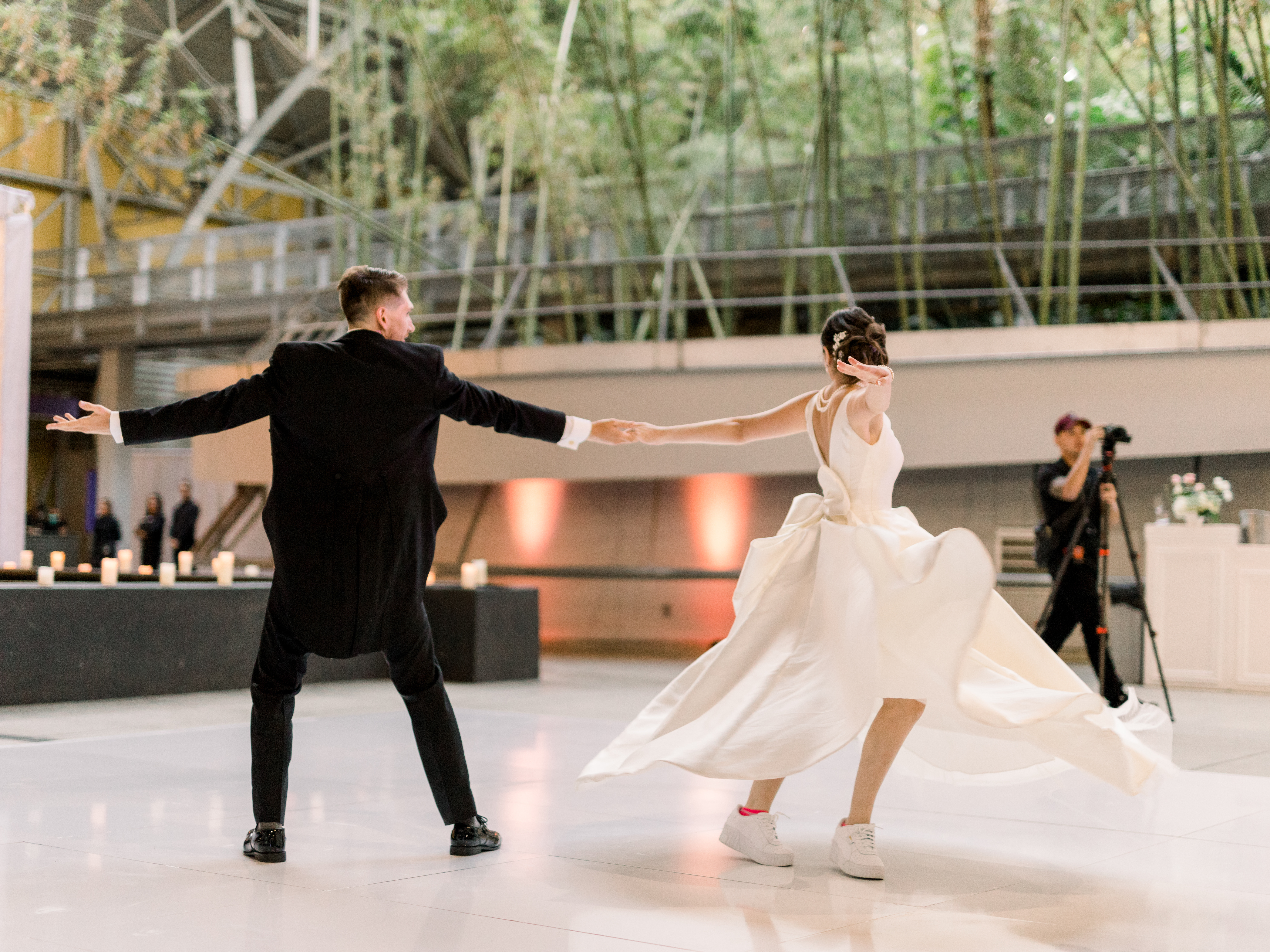 A bride and groom dance on a white dance floor on the east side of the Wallis Annenberg Building: Big Lab. In the background there is a photographer, event lighting, and a bamboo forest.
