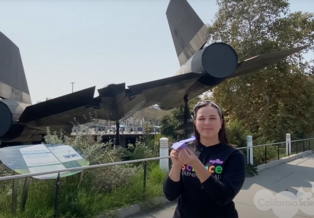 Educator Elaine holds a purple paper plane in front of the Science Center's A-12 trainer aircraft 