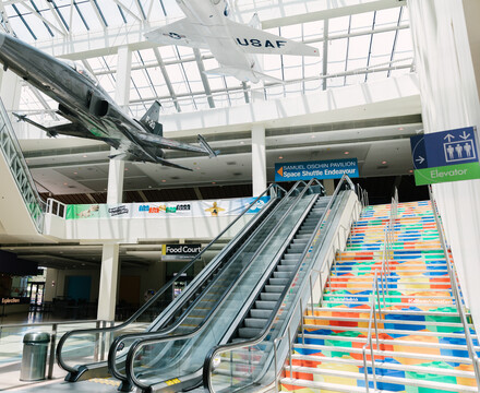 Escalators and stairs leading from Edgerton Court to Disney Science Court. Stairs feature LEGO brick art print on side panels and aircraft are seen displayed overhead
