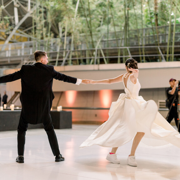 A bride and groom dance on a white dance floor on the east side of the Wallis Annenberg Building: Big Lab. In the background there is a photographer, event lighting, and a bamboo forest.