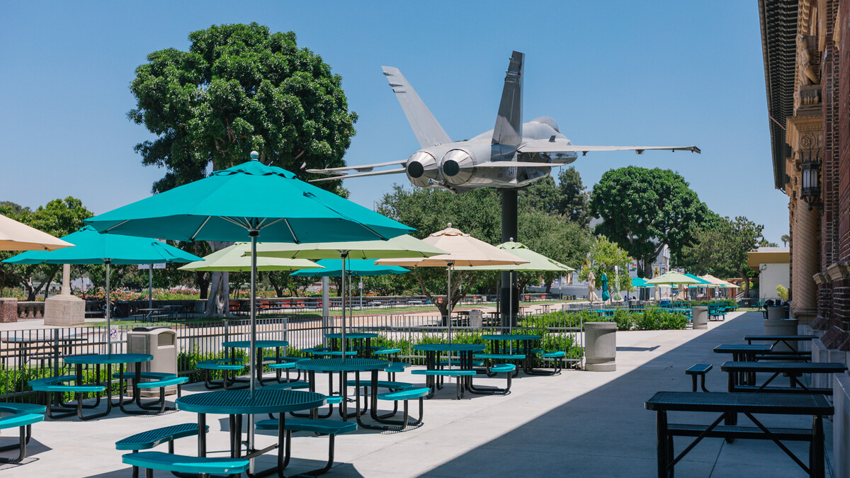 North patios featuring teal picnic benches with umbrellas in pastel yellow and greens, surrounded by lush planters and a tree-lined sidewalk and F/A-18A Hornet between patios
