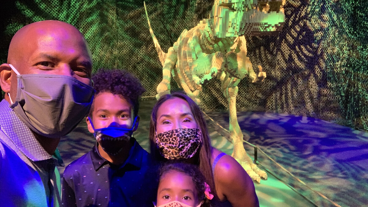 Diverse family with face masks taking a selfie in front of T-Rex sculpture in The Art of the Brick exhibition