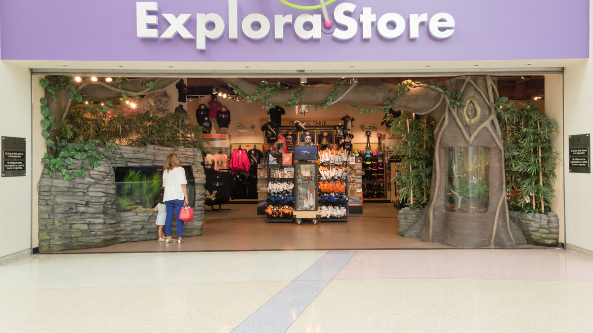 Woman and child stand outside ExploraStore entrance. Upper entry signage reads "Explorastore" and features a large decorative gyroscope. Racks of store merchandise can be seen inside.