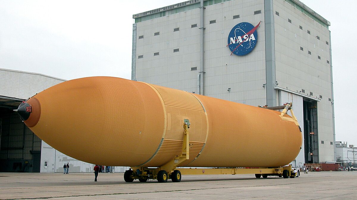 A huge orange external tank lies horizontally on a trailer in front of a large building with a NASA logo on the side.