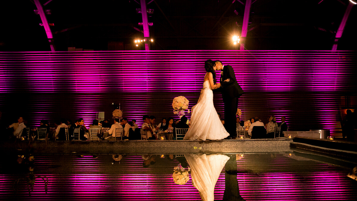 A bride and groom kiss at dusk in front of wedding guests seated at dining rounds and corrugated metal lit in purple. Bride and groom image are reflected in nearby fountain water