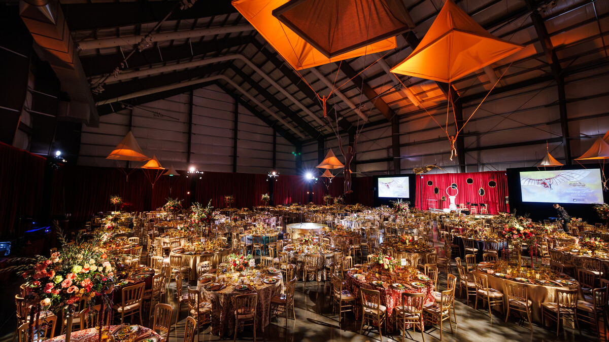 Pavilion hangar space outfitted with perimeter drapery and set with gilded banquet rounds, a central programming stage, and aerial installations