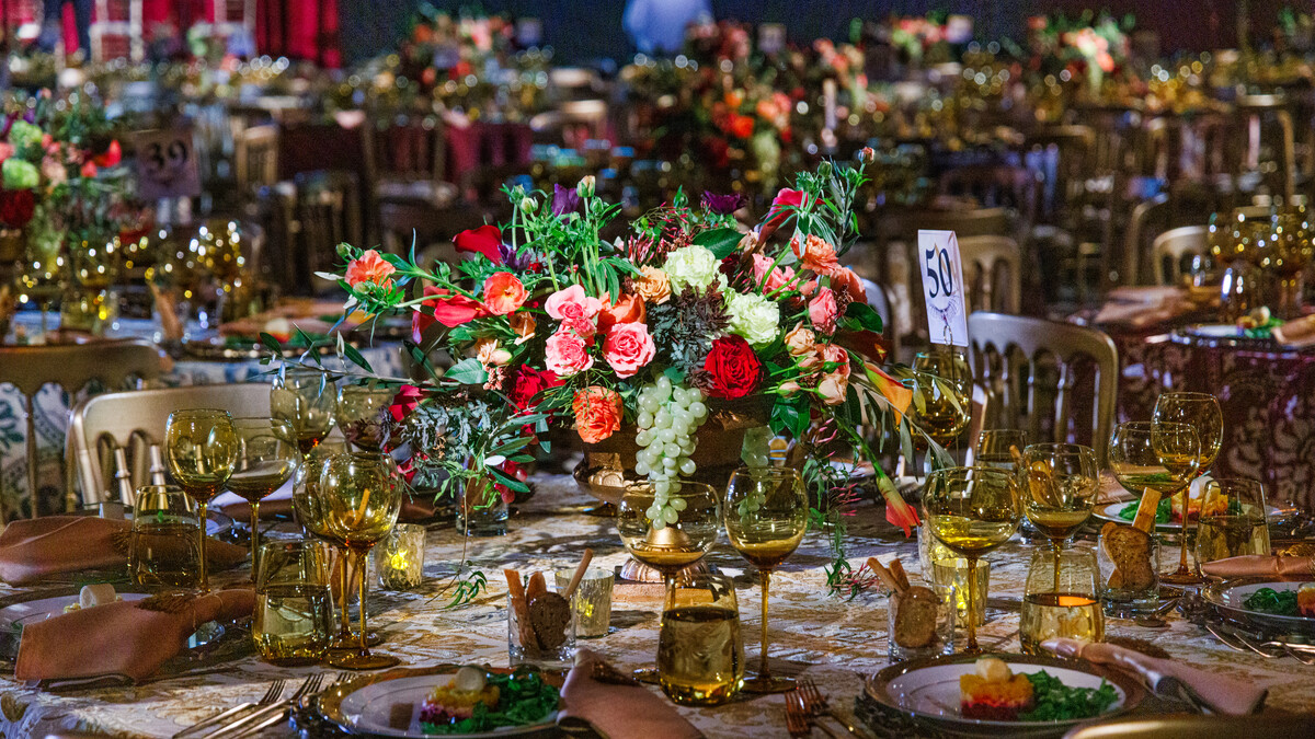 A closeup of a round dinner table with a white-and-gold paisley velvet linen. Each place has a gold chair, a white plate with a gold rim and an appetizer, and golden glasswear. In the center is a pink, red, and green floral arrangements with a hanging cluster of grapes. In the background, slightly blurred, are many similar tables.