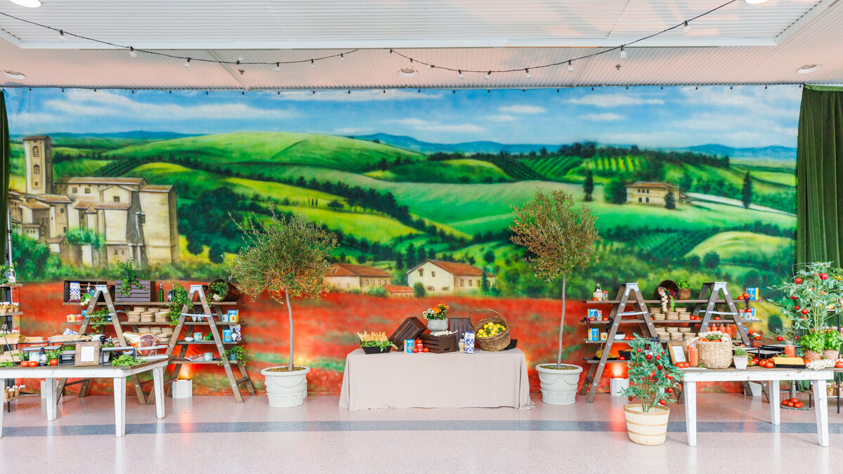 A large painted backdrop of a Tuscan landscape, with green rolling hills, a castle, some small stone houses, and a large red poppy field. In front of it, two mirrored food stations with tables and shelves decorated with potted tomato and basil plants, pasta, canned tomatoes, and baskets of fruits and vegetables. 