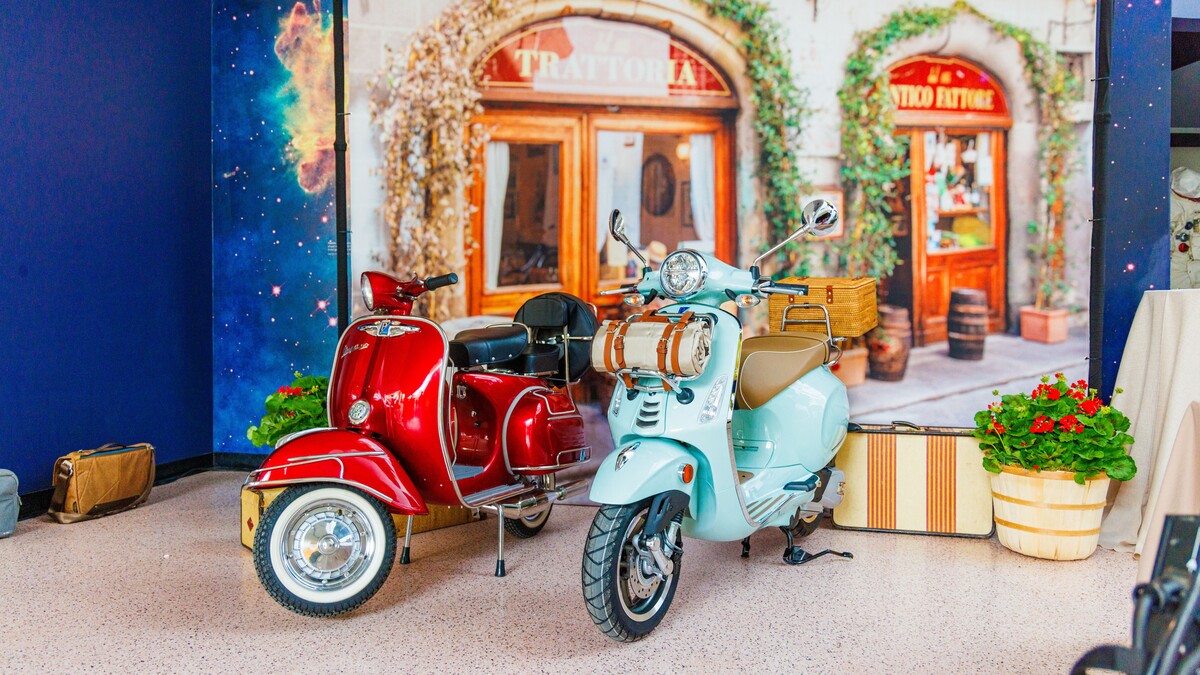 A photo booth setup; a backdrop of a slightly blurred Italian city scene with two arched doorways. In front of that, two Vespa scooters; a red one to the left and a mint green one to the right. To their right is a prop vintage suitcase and a potted pink geranium plant.
