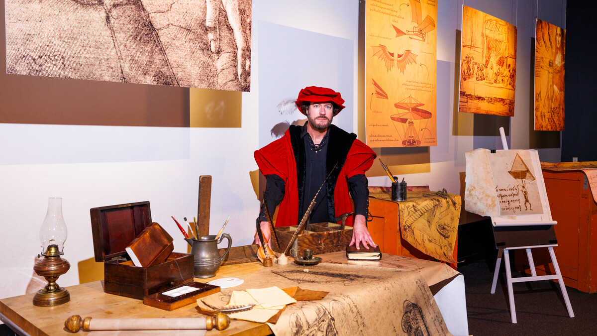 A man with a beard, dressed as Leonardo da Vinci in black and red robes and a red feathered cap, stands behind a desk covered in historical props: parchment, drawn diagrams, an oil lamp, a painters box with brushes, quills, inkwells, etc. Behind him, suspended da Vinci drawings and diagrams and an easel with a propped drawing of an early parachute design.
