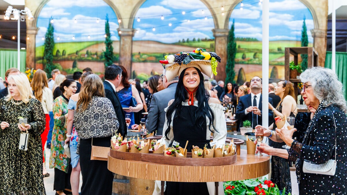 A cocktail party. In the foreground, a woman with long black hair and a fruit basket hat stands in a circular table with cups of charcuterie meats and cheeses. Around here, guests in formalwear mingle and eat. In the background, a painted backdrop of a hilly Tuscan landscape, framed by three stone arches and four real potted cypress trees.