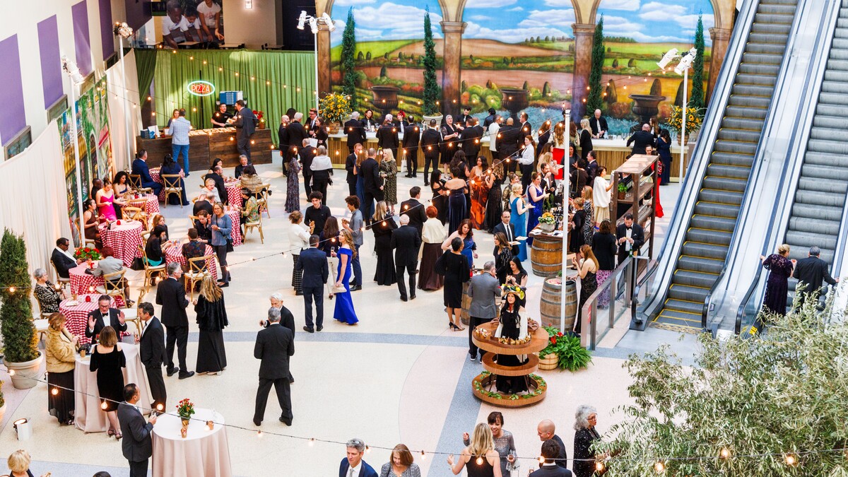 An aerial view of Edgerton Court at the California Science center, filled with guests in formalwear. String lights criss-cross over the party, there are tall cocktail tables and short round tables with red-and-white checkered tablecloths. In the background, a painted backdrop of a hilly Tuscan landscaped framed by 3 stone arches, and flanked by 4 real potted cypress trees.