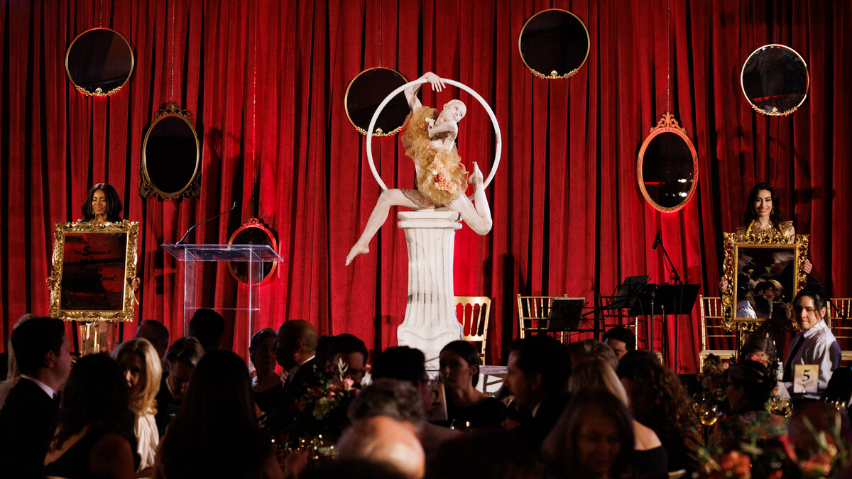 In the foreground, a dimly lit crowd of party guests sit at round tables, chatting and eating. Behind them, a stage with a red-draped back wall, decorated with suspended mirrors in gold frames. On the center of the stage is a white stone column with a hula-hoop shaped ring on top, in which sits a contortionist performer painted all white in a renaissance outfit.
