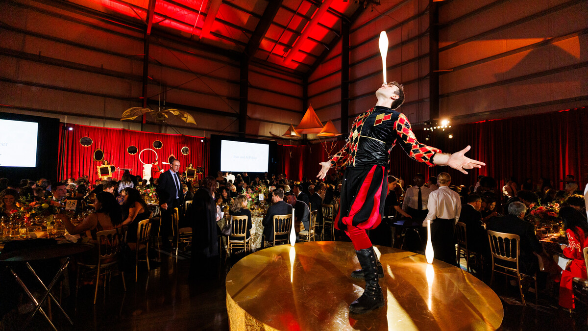 A dinner party. In the background, dimly lit party guests in formal wear sit at round tables in gold chairs. In the distance, a red velvet draped wall and a hanging da Vinci flying machine. In the foreground, on a small gold stage, a juggler in a jester outfit balances a juggling club on his nose.